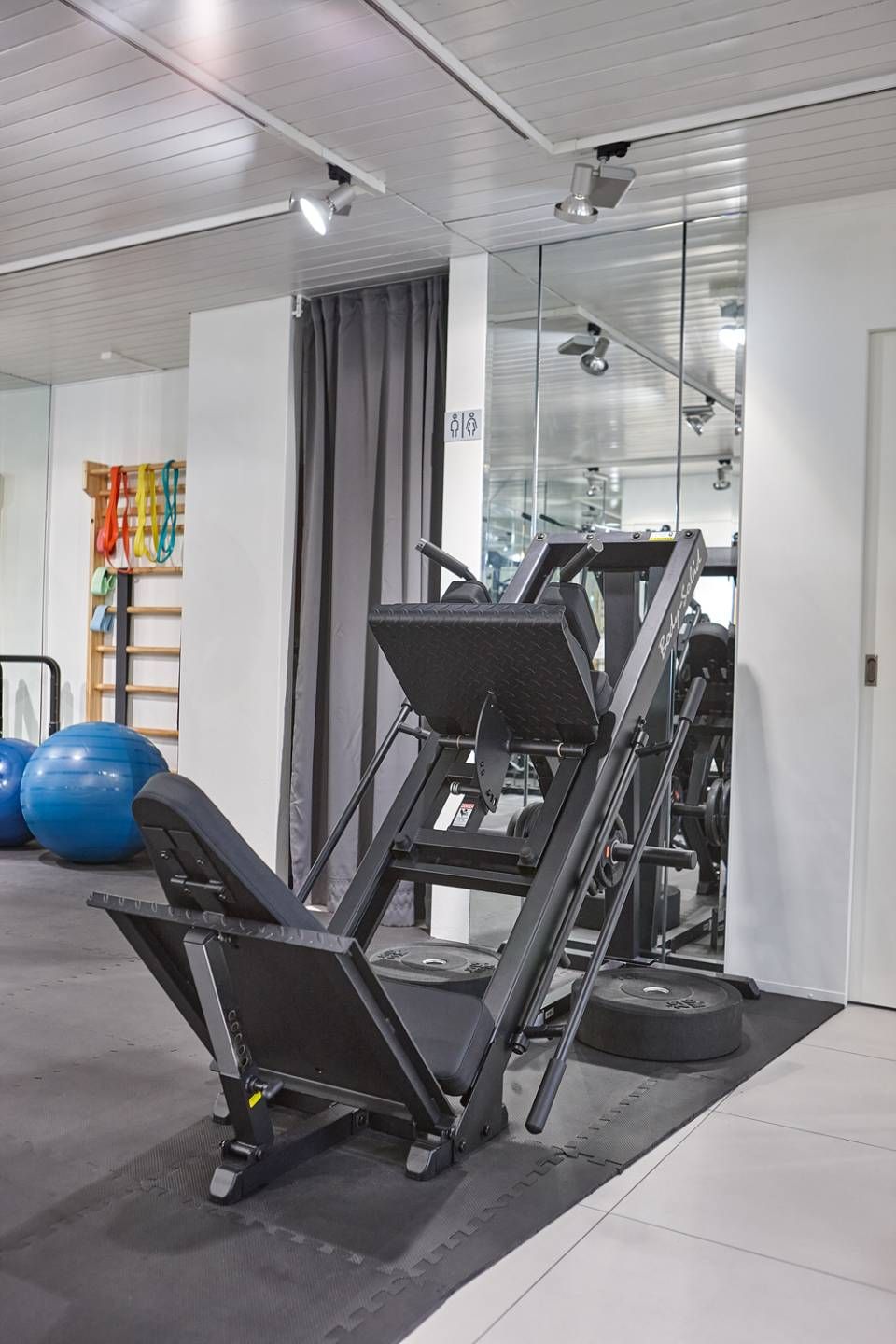In einem Fitnessstudio steht vor einem Spiegel eine Beinpresse, daneben befinden sich ein blauer Gymnastikball und an der Wand montierte Geräte.