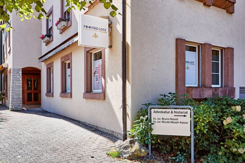 Ein Arzt in einem blauen Hemd bespricht das bandagierte Bein eines Patienten, das in einer Arztpraxis auf einem Hocker ruht.