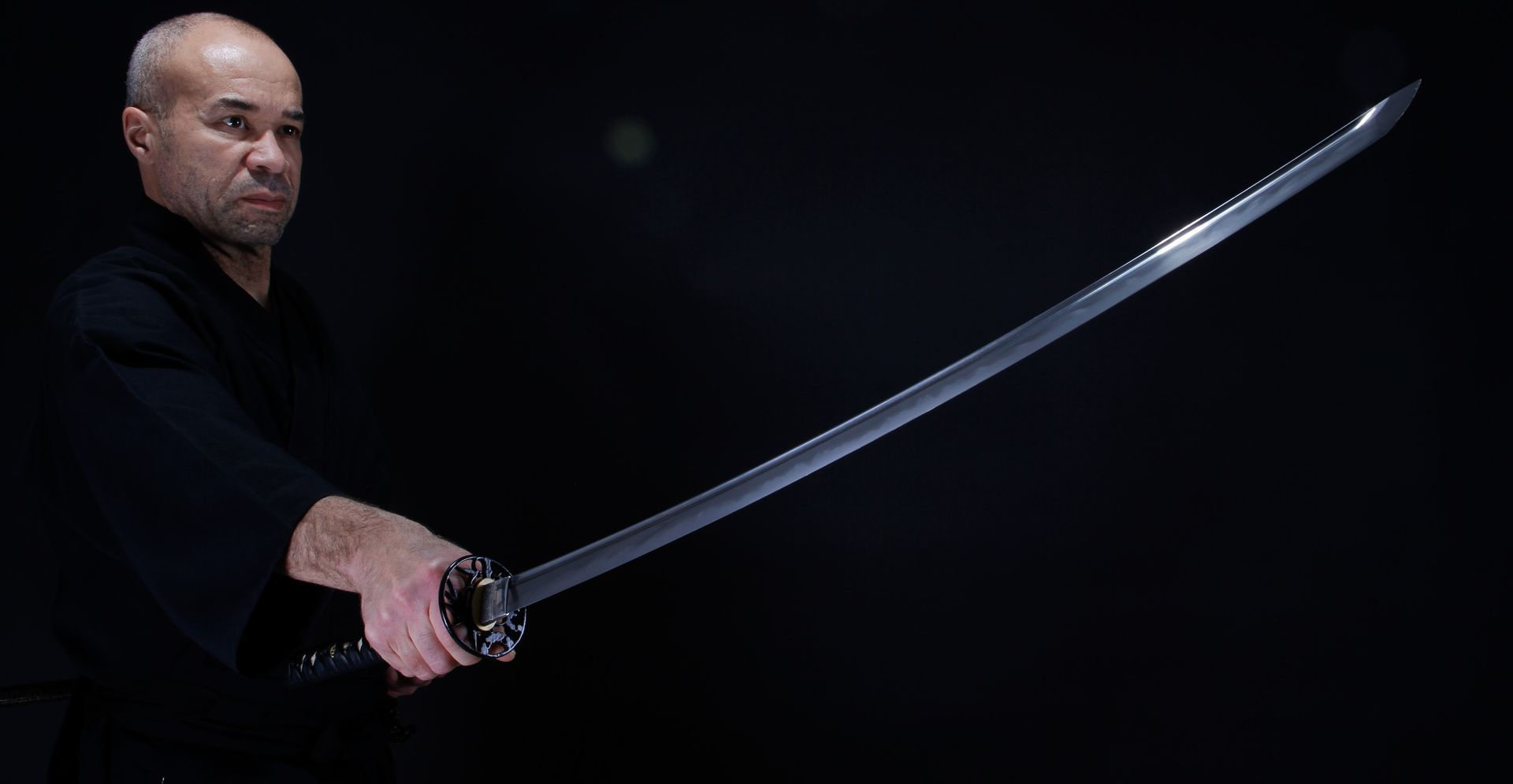 Un homme vêtu de noir tient un katana, la lame étincelante, sur fond sombre.