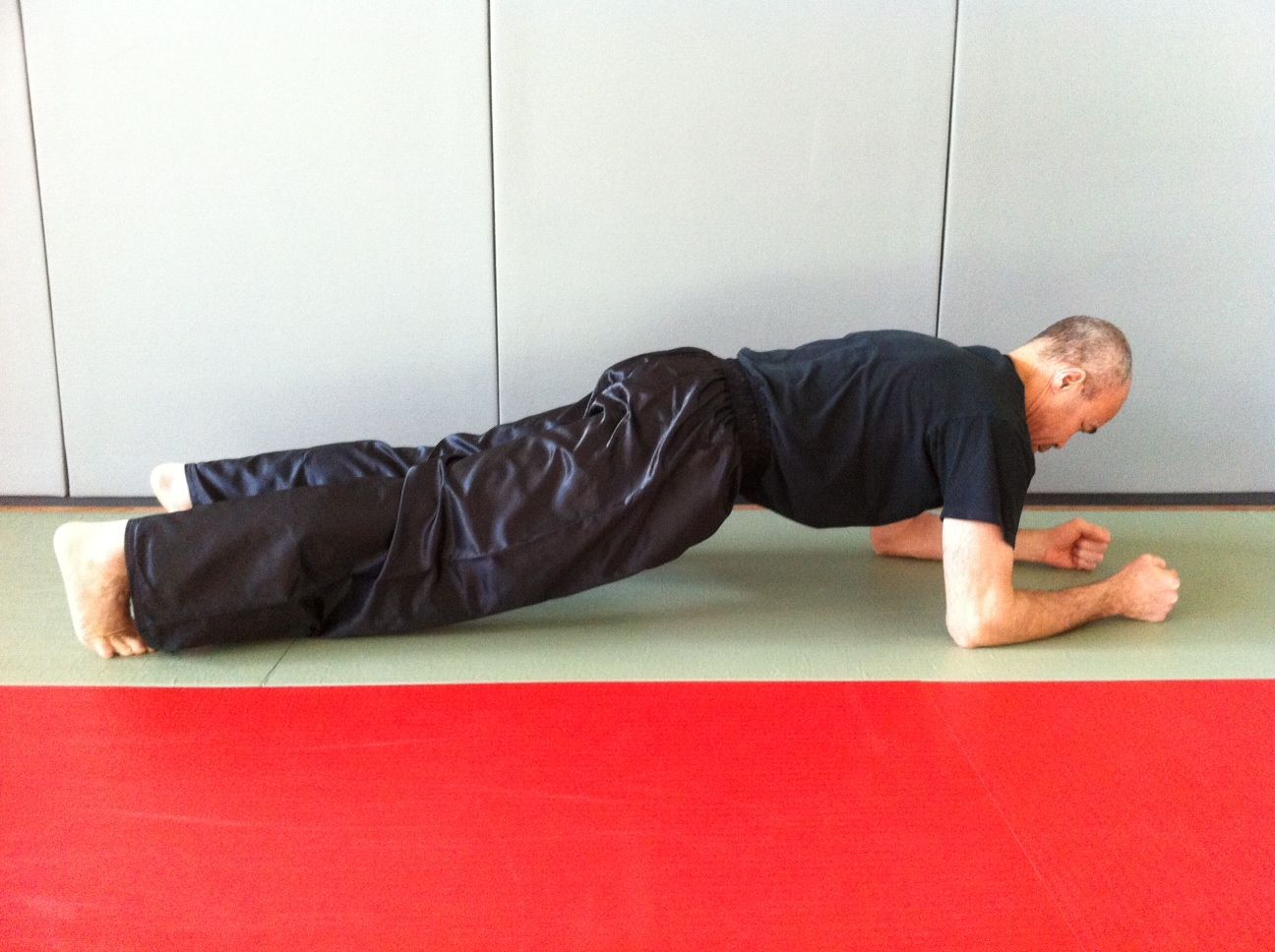 Un homme vêtu de noir est en position de planche latérale sur un tapis rouge et vert. Sa main gauche est levée.