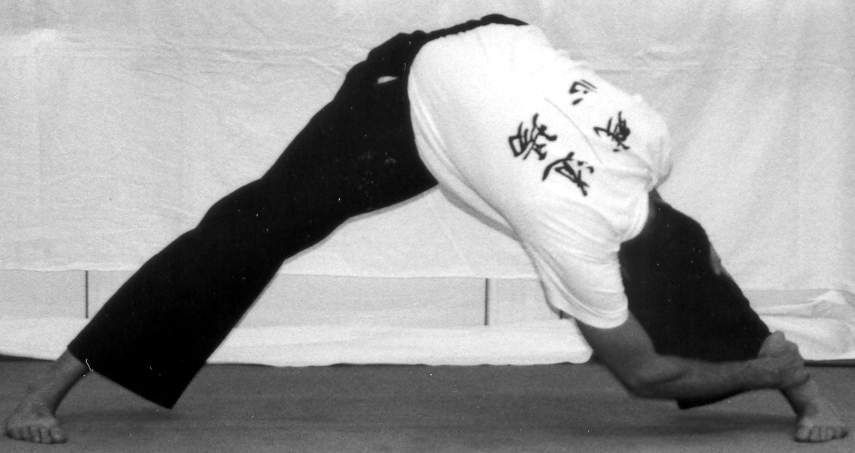 Un homme en chemise blanche et pantalon noir effectue une posture de yoga, les jambes écartées, penché en avant.