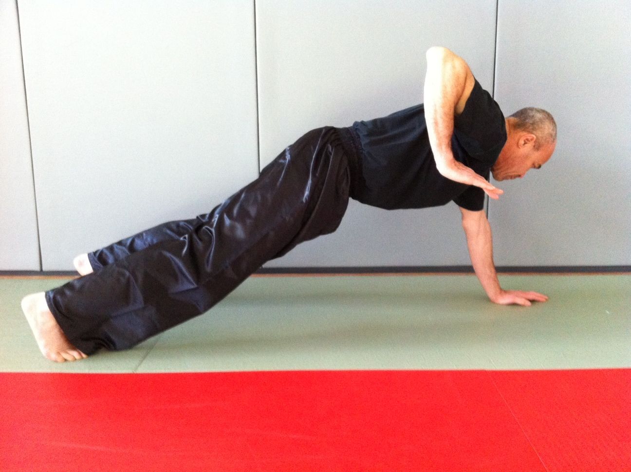 Un homme fait une pompe, un bras tendu, sur un tapis rouge et vert contre un mur gris.