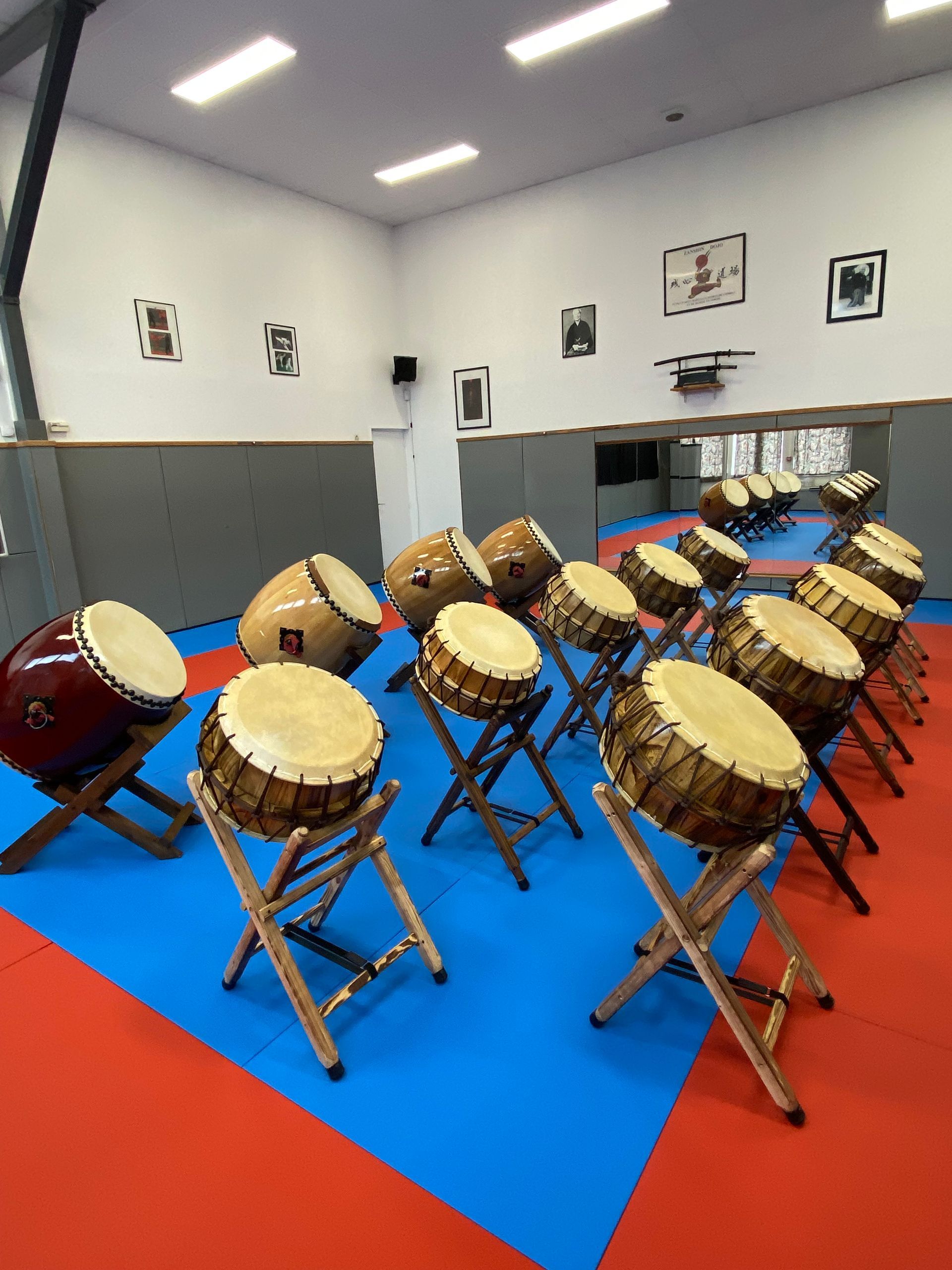 Rangée de tambours taiko dans un dojo, placés sur des supports en bois au-dessus d'un tapis bleu et rouge.