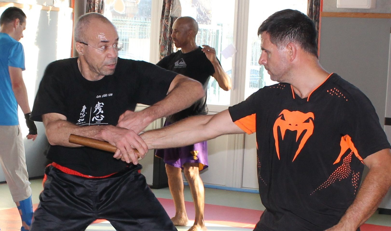 Deux hommes pratiquent des techniques de défense au bâton dans un cours d'arts martiaux. D'autres personnes sont visibles en arrière-plan.