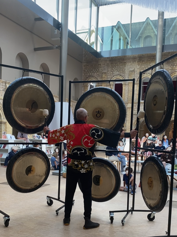 Une personne joue de plusieurs gongs dans un intérieur. Elle porte une veste colorée. Les gongs sont placés sur des supports à roulettes.