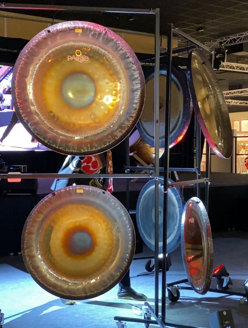 Des gongs sur un support métallique, de tailles variées, dans des tons d'or et de bleu. Une personne se tient à proximité.