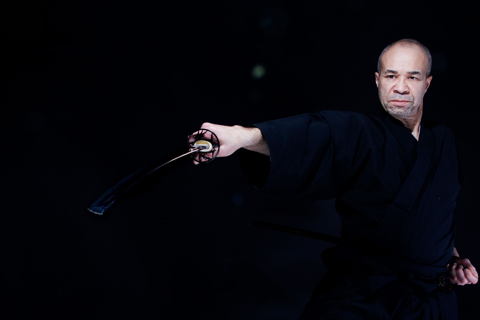 Homme en kimono noir, brandissant un katana, devant un fond sombre.