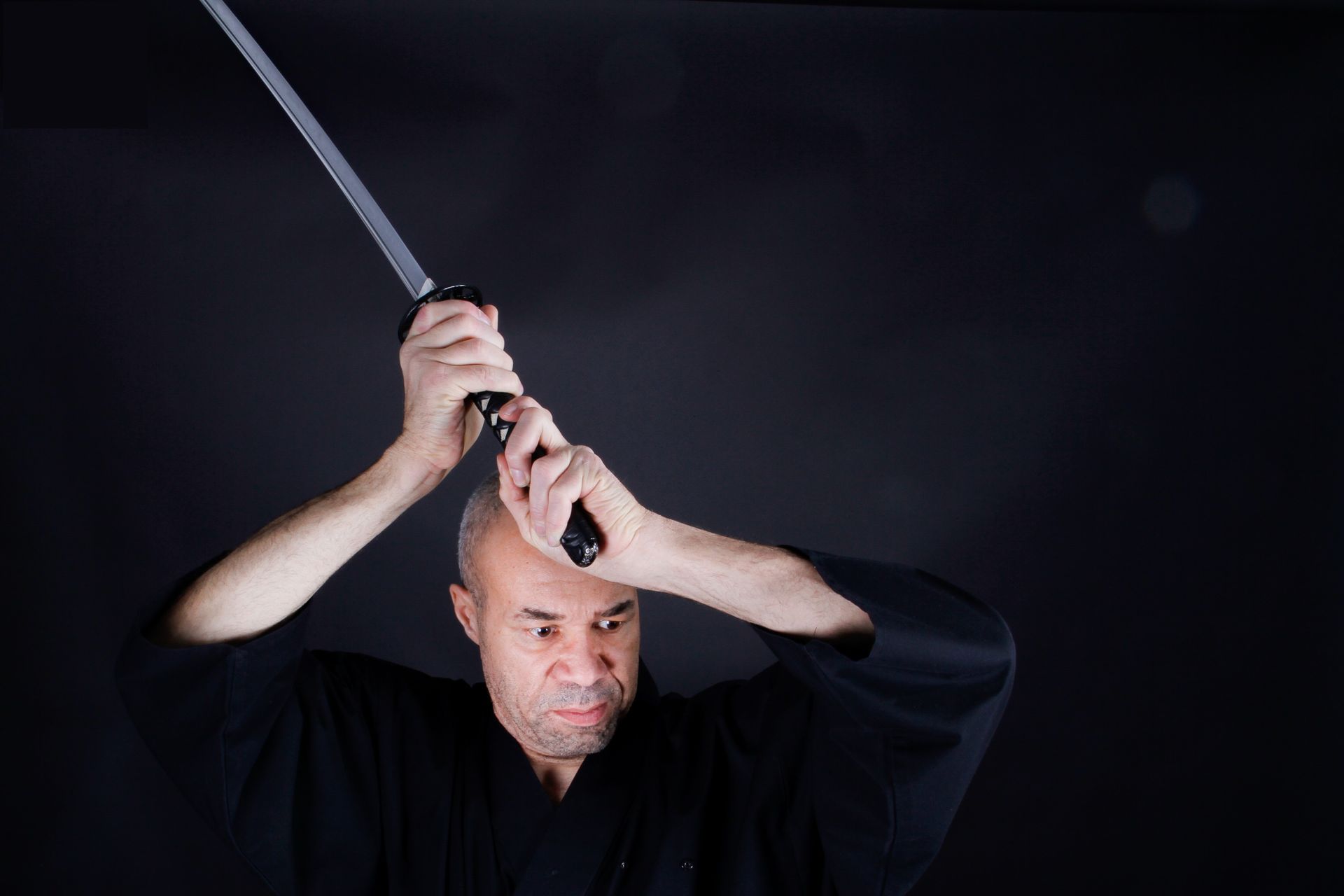 Un homme en robe noire, brandissant un katana, exécute une figure d'arts martiaux. Fond sombre.