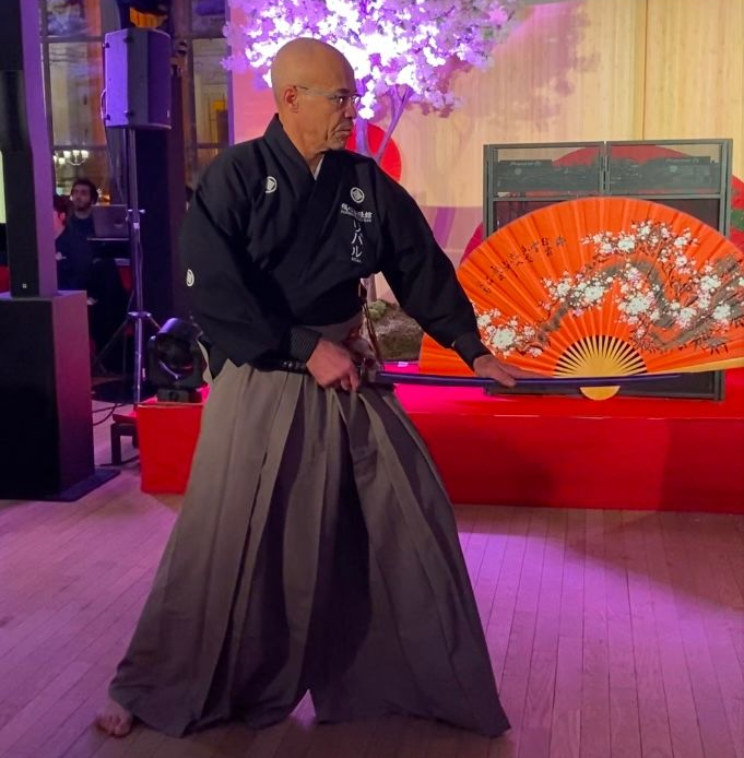 Un homme en tenue japonaise, tenant un éventail et un katana, exécute une performance. Décor de cerisiers en fleurs, scène.