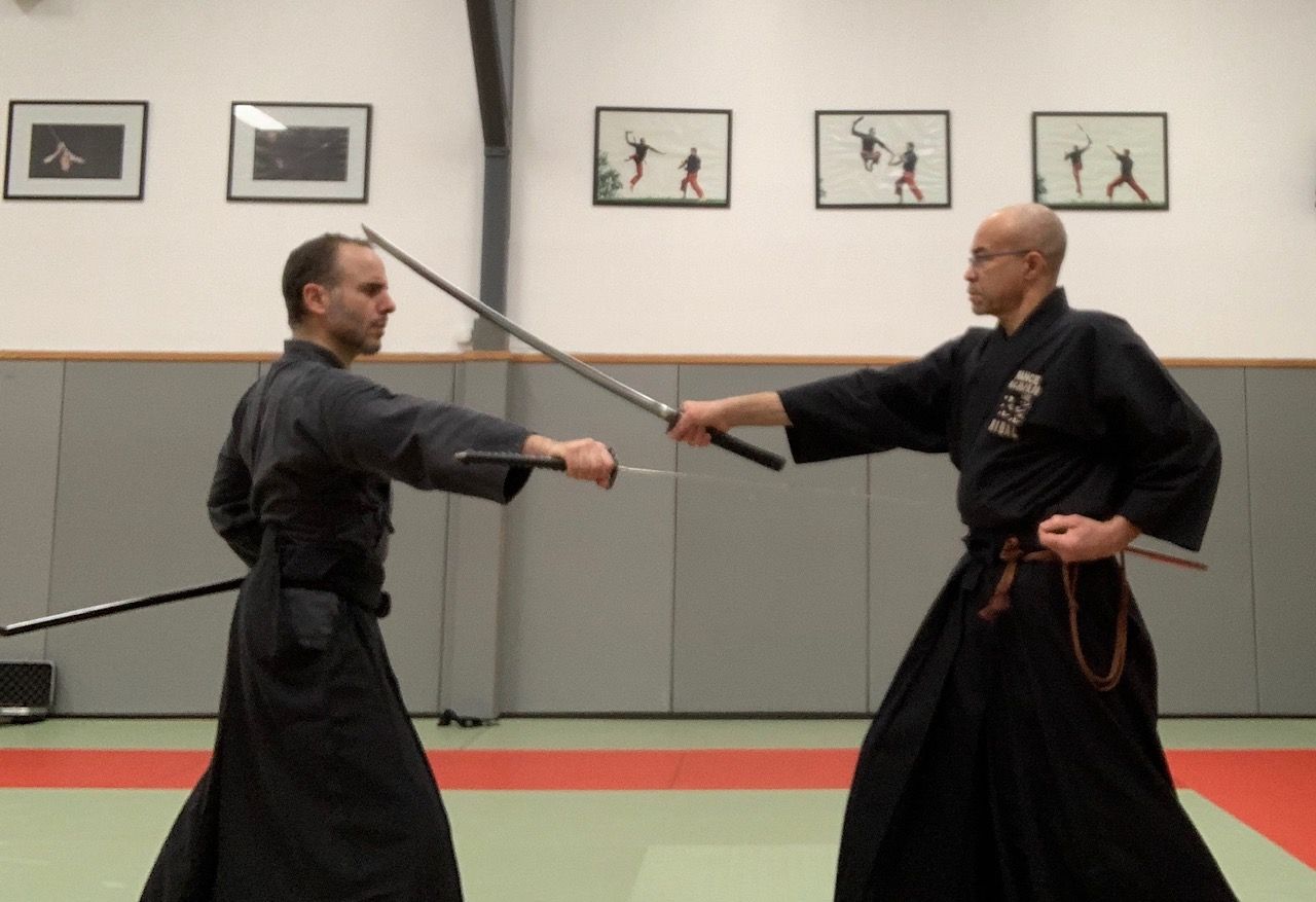 Deux personnes en tenue d'arts martiaux noire s'entraînent au combat à l'épée dans un dojo.