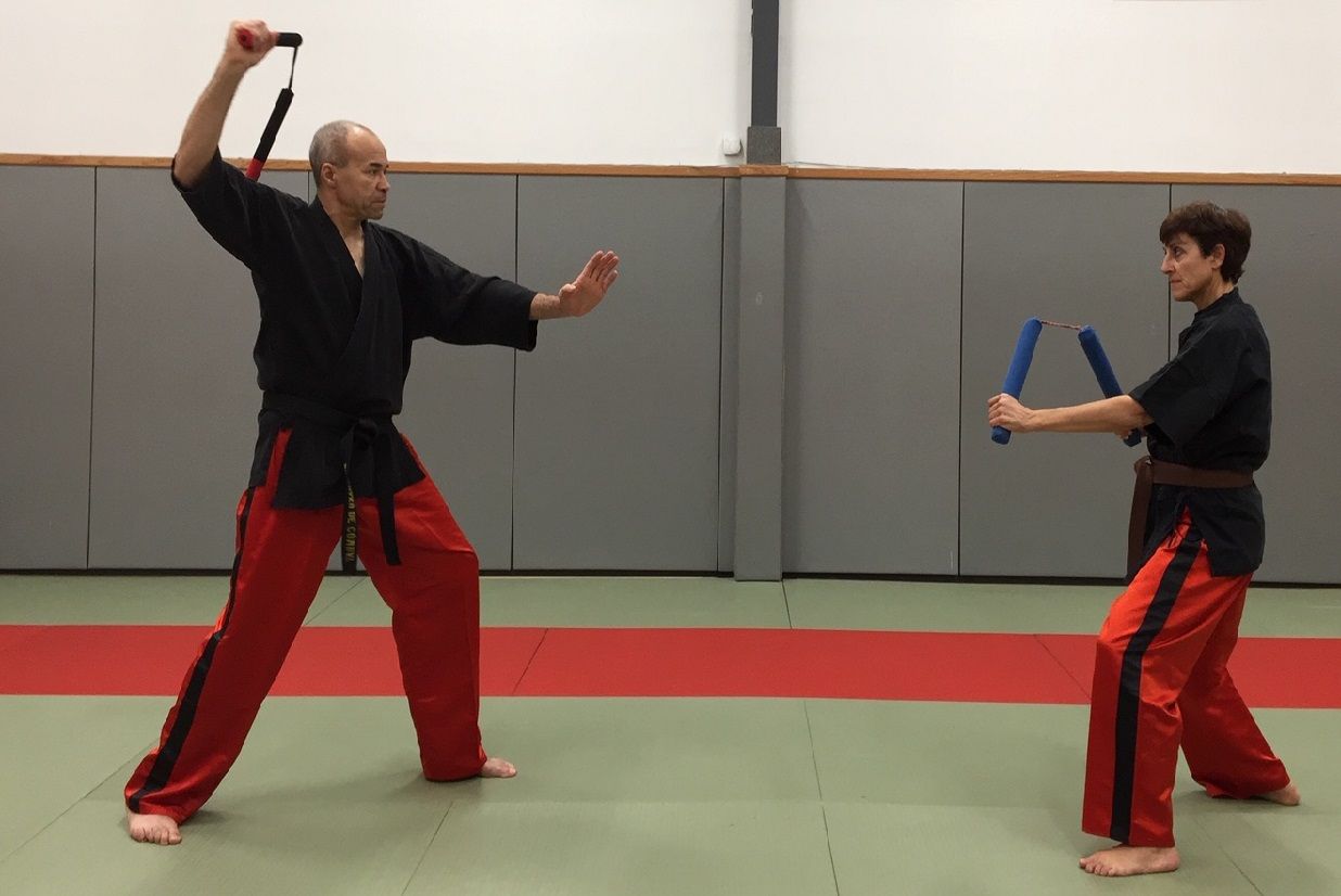 Deux personnes pratiquent les arts martiaux. L'une manie le nunchaku, l'autre le tient, toutes deux vêtues de pantalons rouges et de hauts noirs.