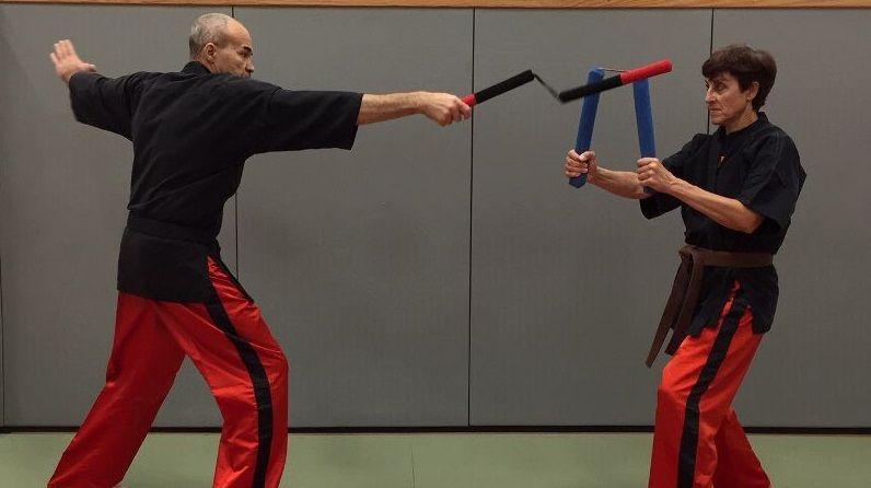 Deux personnes pratiquent les arts martiaux avec des armes. L'une frappe avec un long bâton, l'autre se défend avec des nunchakus.