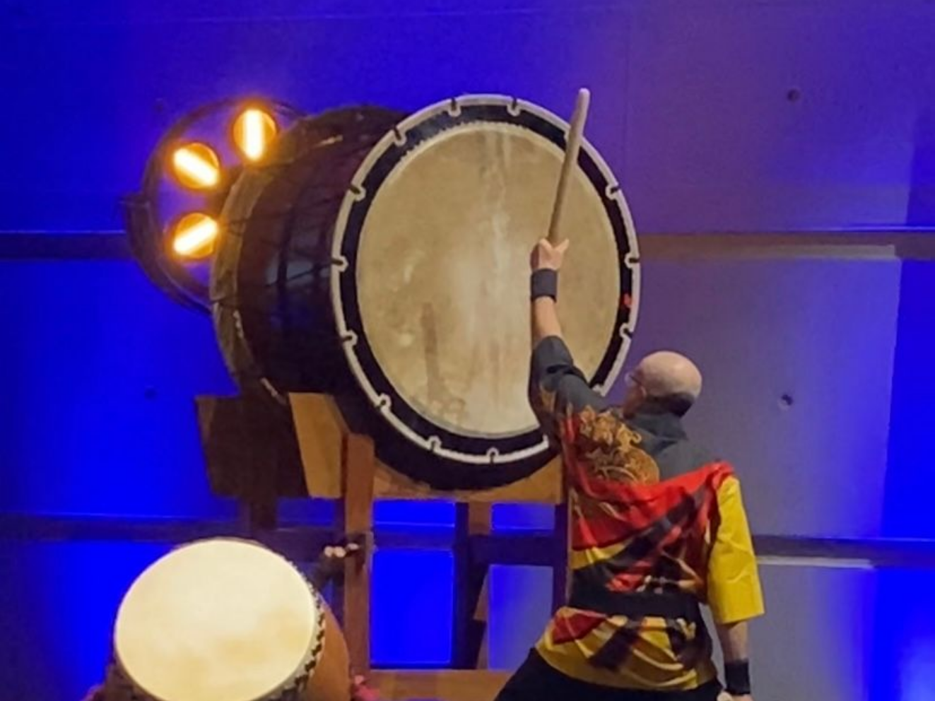 Une personne joue du taiko, un grand tambour avec une baguette. La scène est éclairée en bleu et orange.