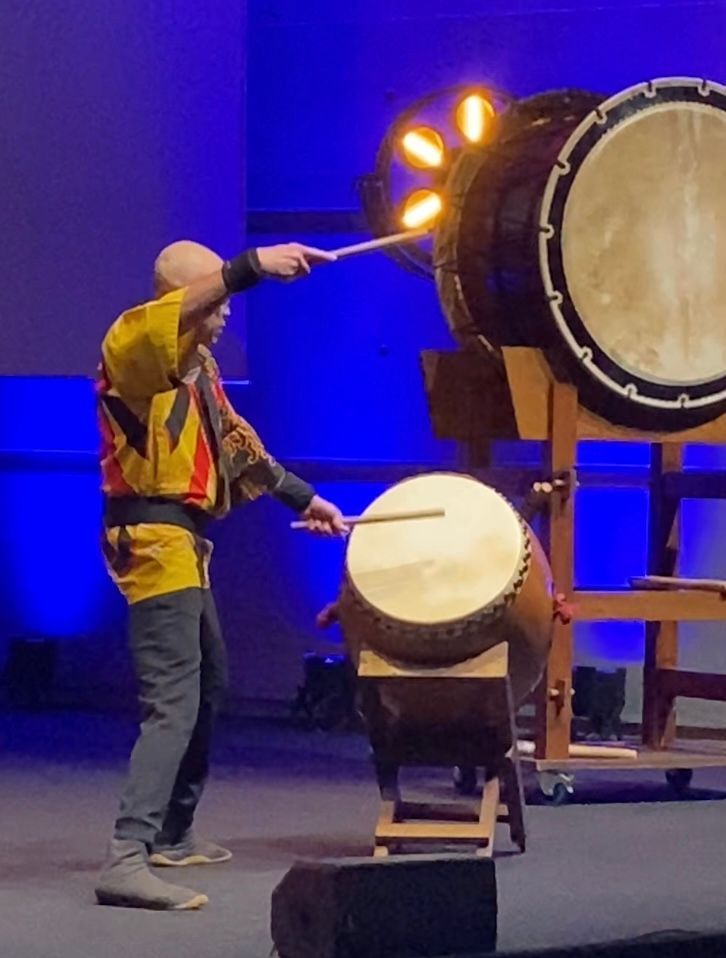 Une personne chauve vêtue d'une veste colorée joue du taiko, un grand tambour, avec des baguettes sur scène, baignée de lumière bleue.