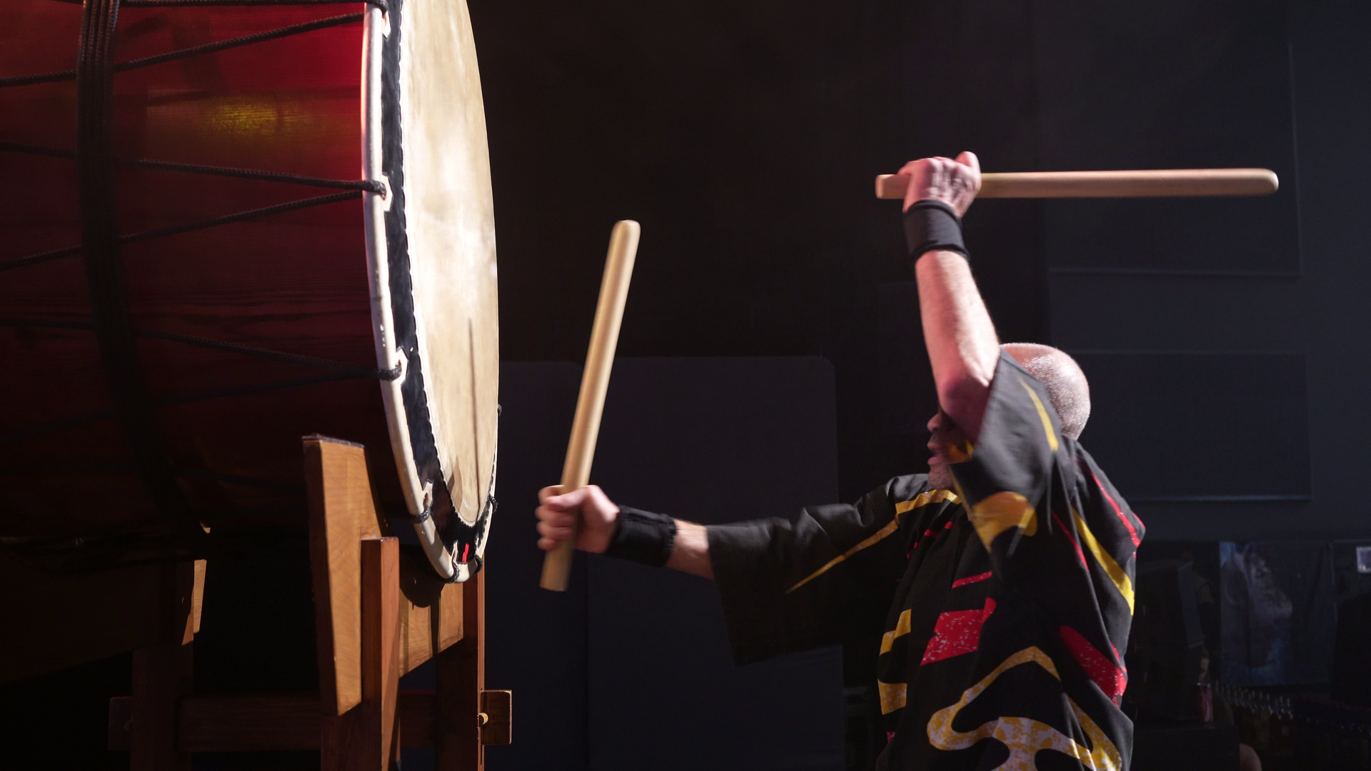 Personne jouant d'un grand tambour taiko avec des baguettes, à l'intérieur.