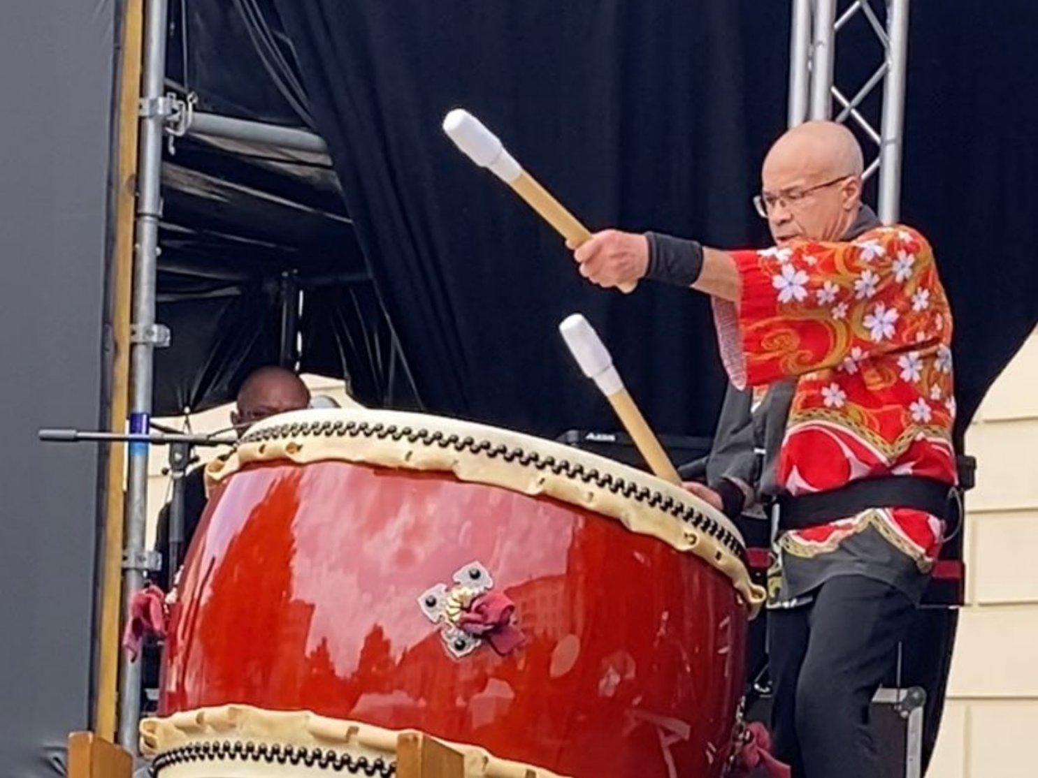 Une personne joue du tambour taiko rouge avec deux baguettes à embouts blancs en extérieur. Elle porte une robe rouge à motifs.