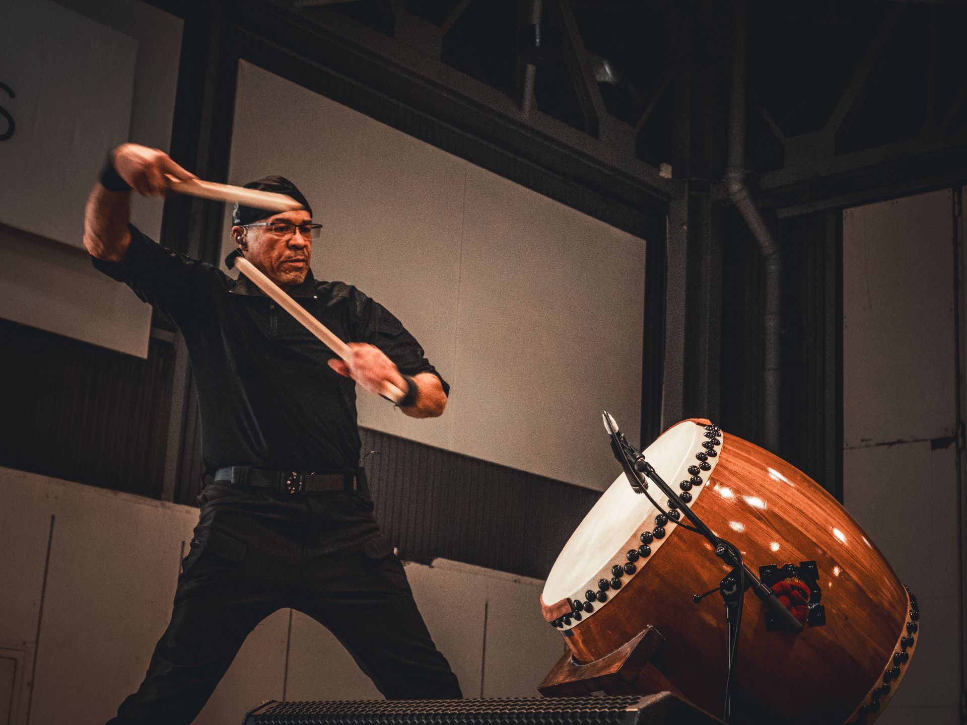 Un homme joue d'un grand tambour taiko sur scène, le frappant avec des baguettes. Vêtements noirs, tambour en bois.