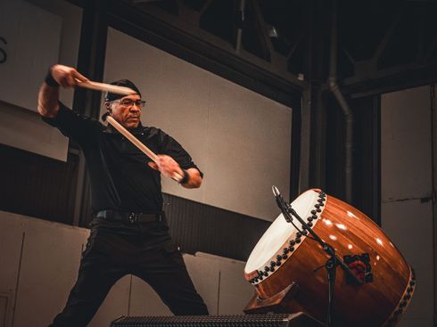 Un homme joue d'un grand tambour taiko sur scène, le frappant avec des baguettes. Vêtements noirs, tambour en bois.
