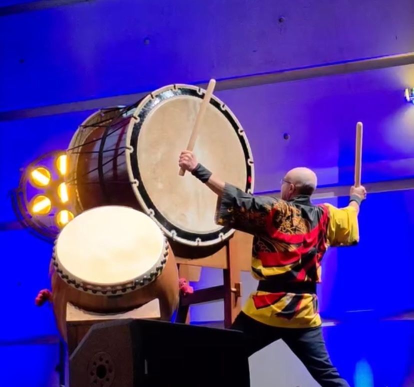 Un batteur jouant de grands tambours taiko avec des maillets en bois sur scène, sur fond bleu.