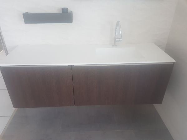 Tocador de pared marrón con encimera y lavabo blancos, grifo plateado y un estante negro sobre una pared blanca.