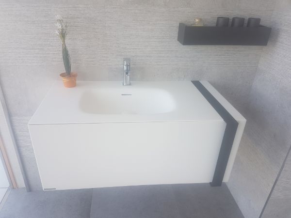 Lavabo de baño flotante blanco con grifo cromado, estante negro y una pequeña planta en maceta contra una pared de azulejos grises.
