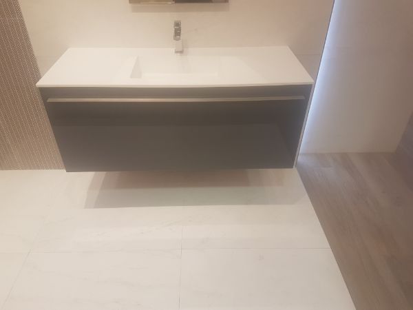 Tocador de baño flotante gris con encimera y lavabo blancos, grifo cromado, sobre suelo de baldosas blancas.