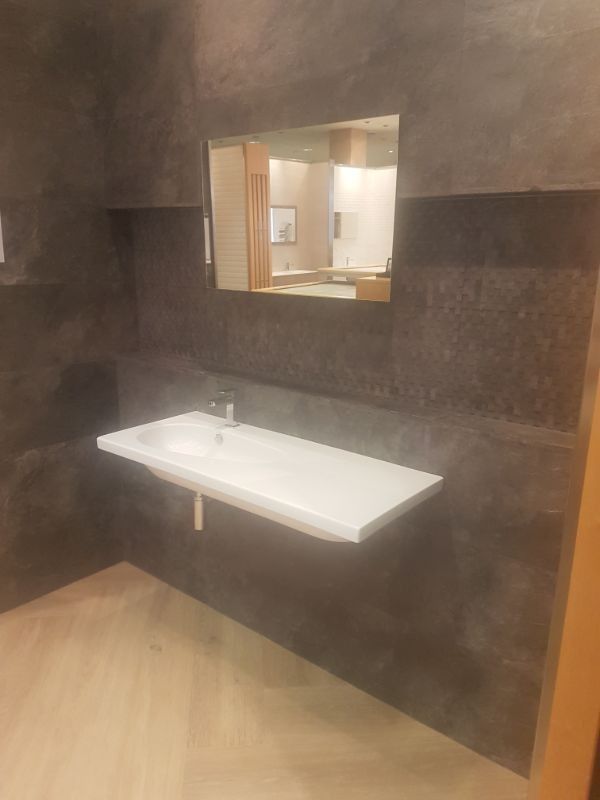 Baño con lavabo blanco montado en la pared, espejo y paredes revestidas de color gris oscuro.