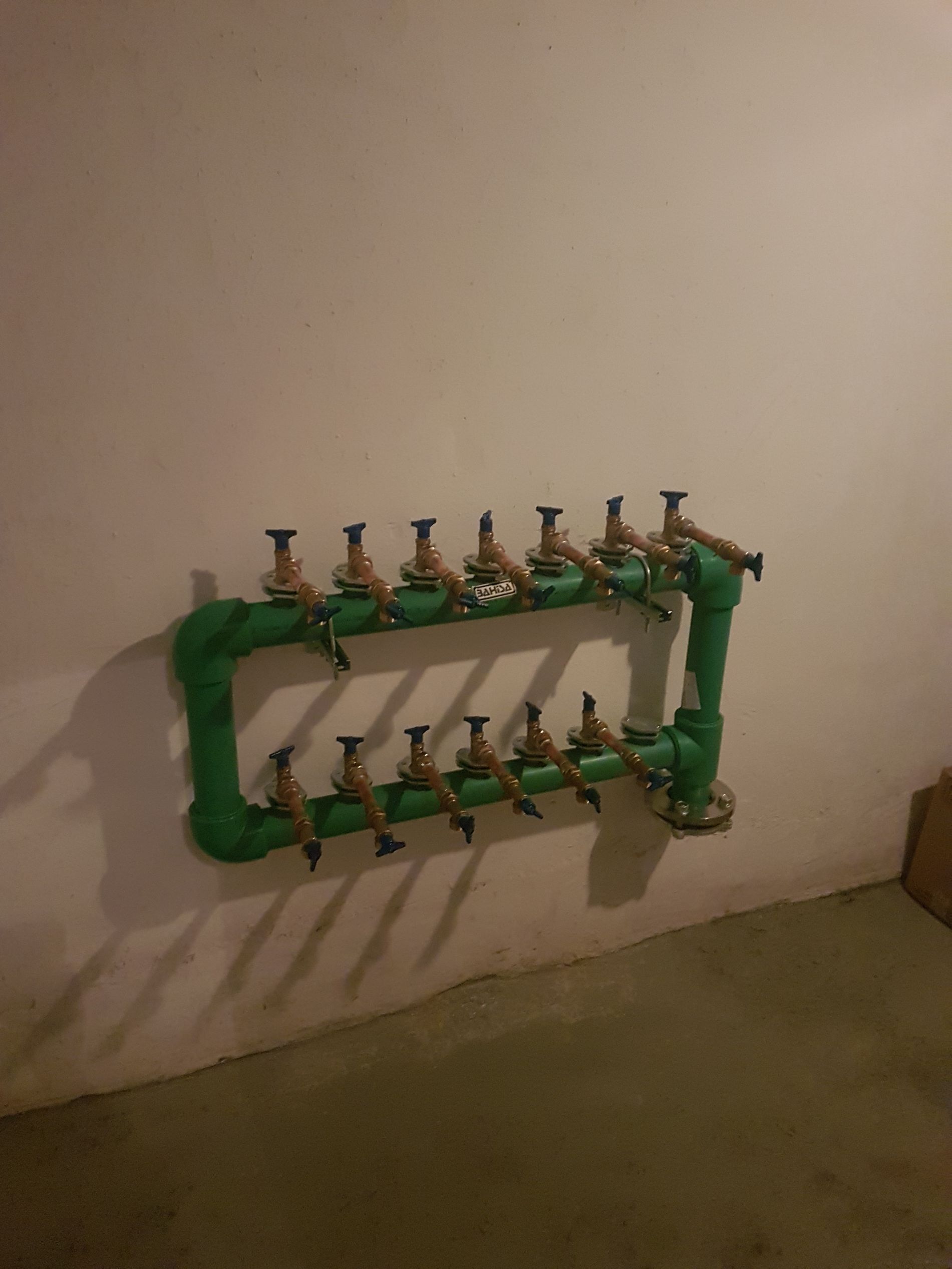 Colector de plomería verde con múltiples válvulas montado en una pared blanca en una habitación con piso gris.