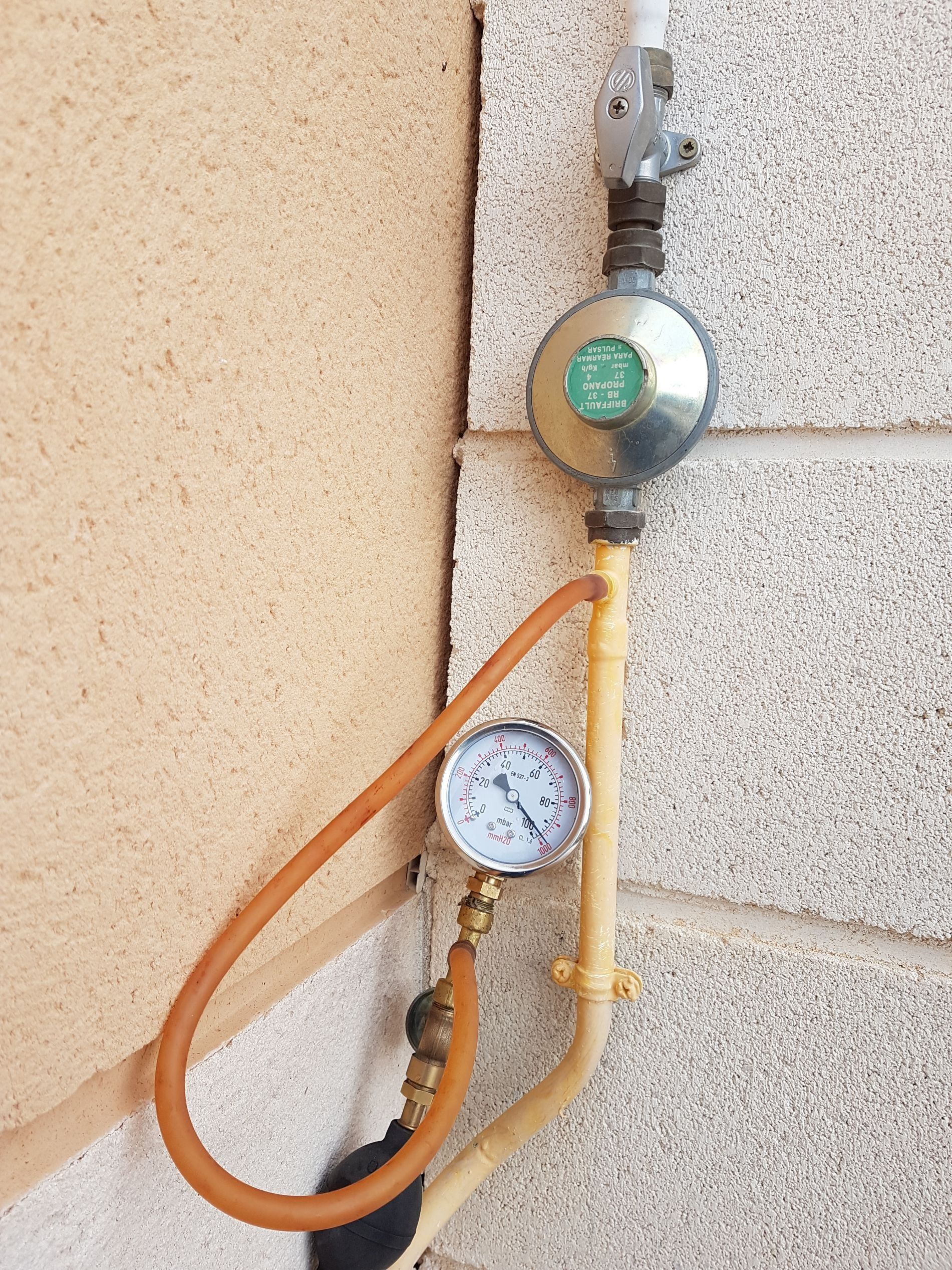 Regulador y medidor de gas en una pared exterior texturizada, conectados mediante mangueras.