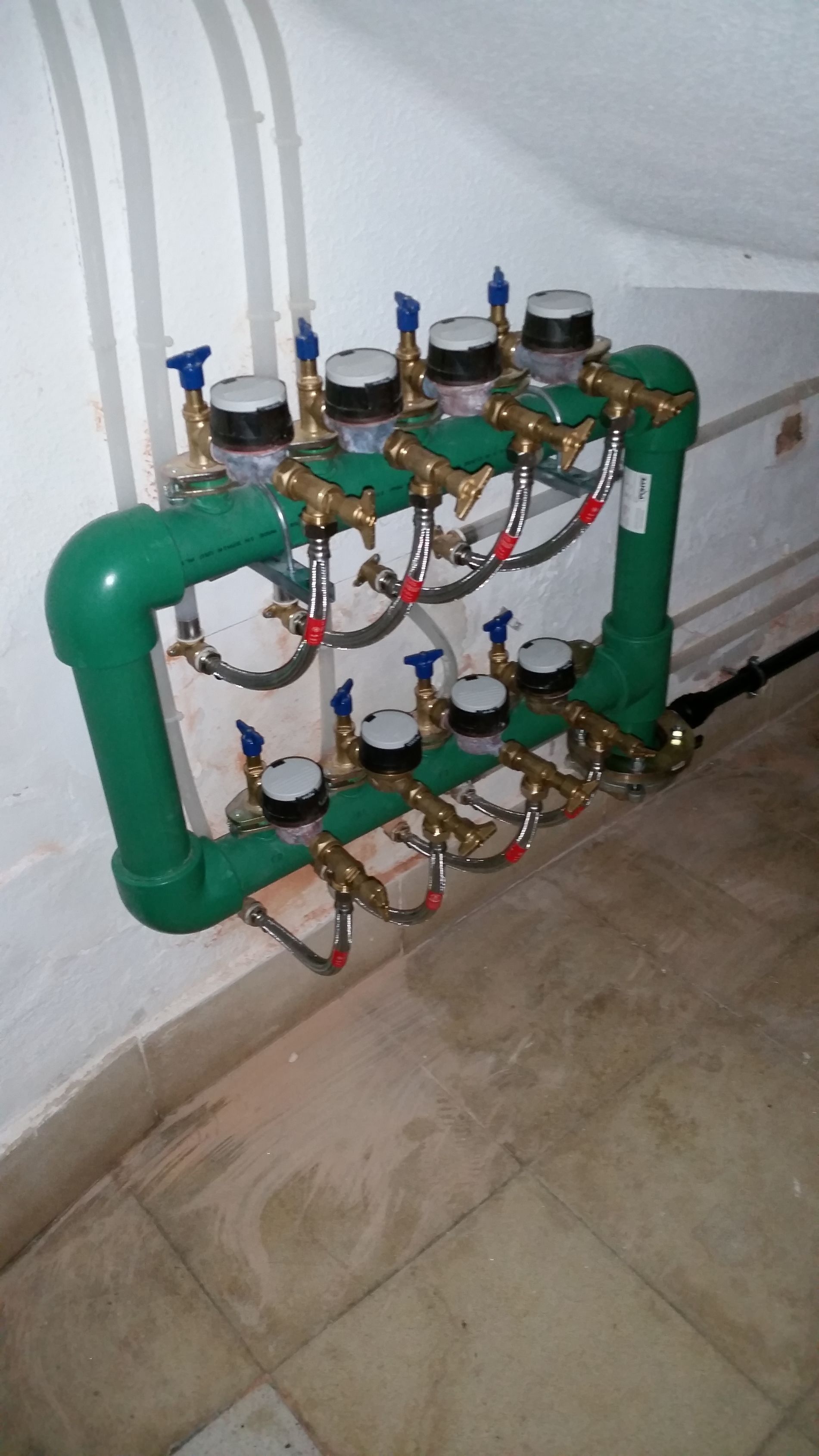 Sistema de medidor de agua verde y latón adosado a una pared blanca en una habitación con suelo de baldosas.