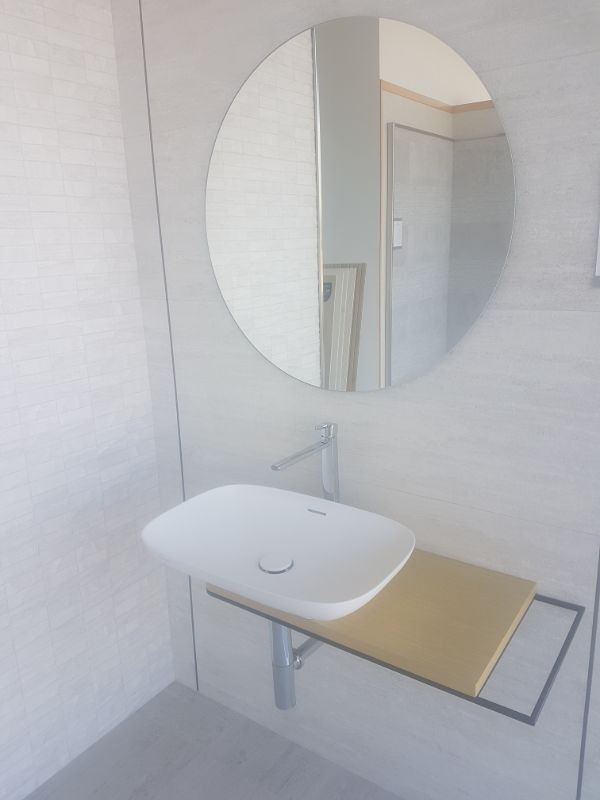 Lavabo de baño moderno con espejo redondo, paredes blancas y estante en tono madera.