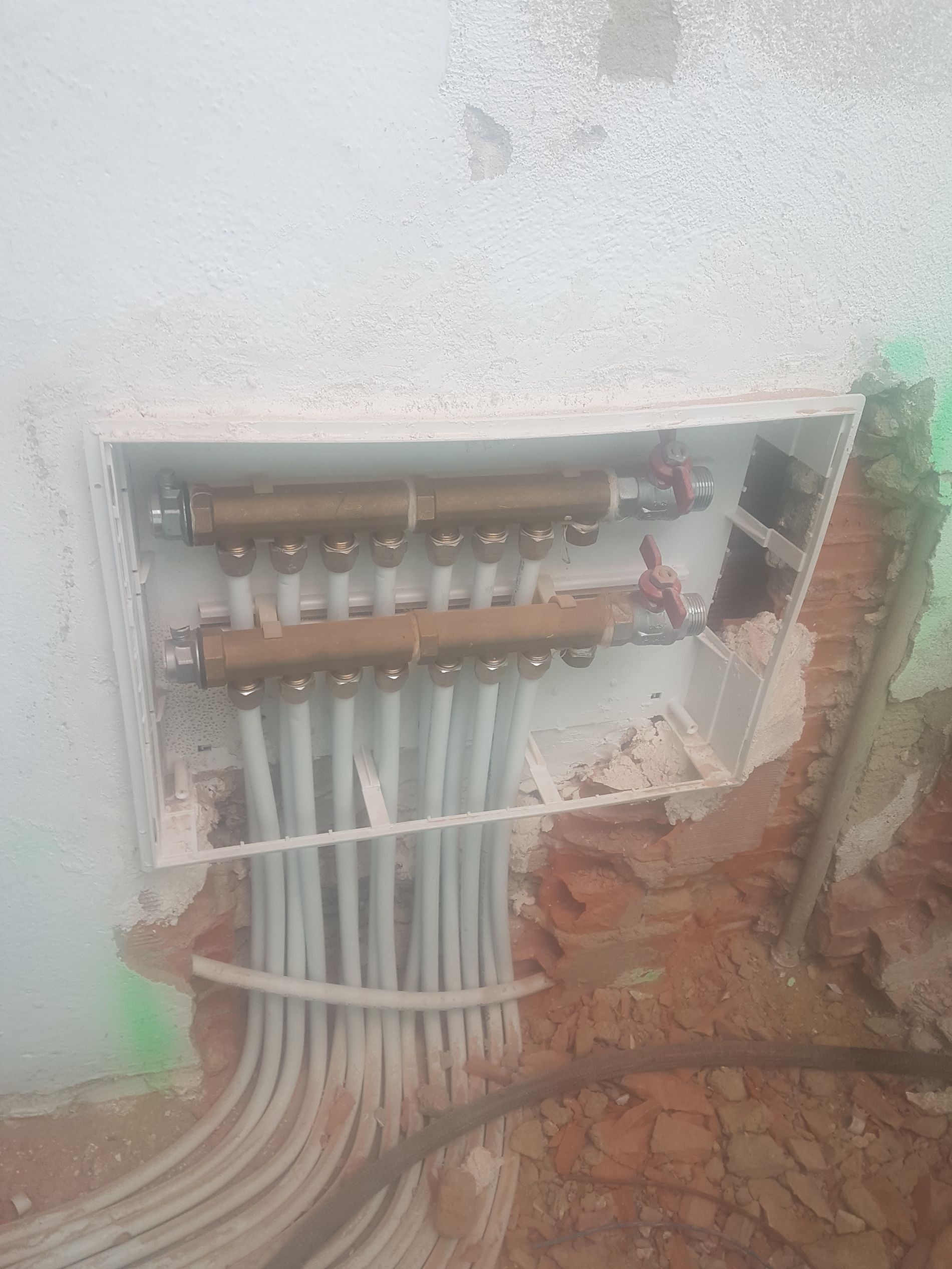 Caja colectora de plomería de color blanco empotrada en la pared, que contiene tuberías de cobre y tuberías grises que se extienden hacia abajo.