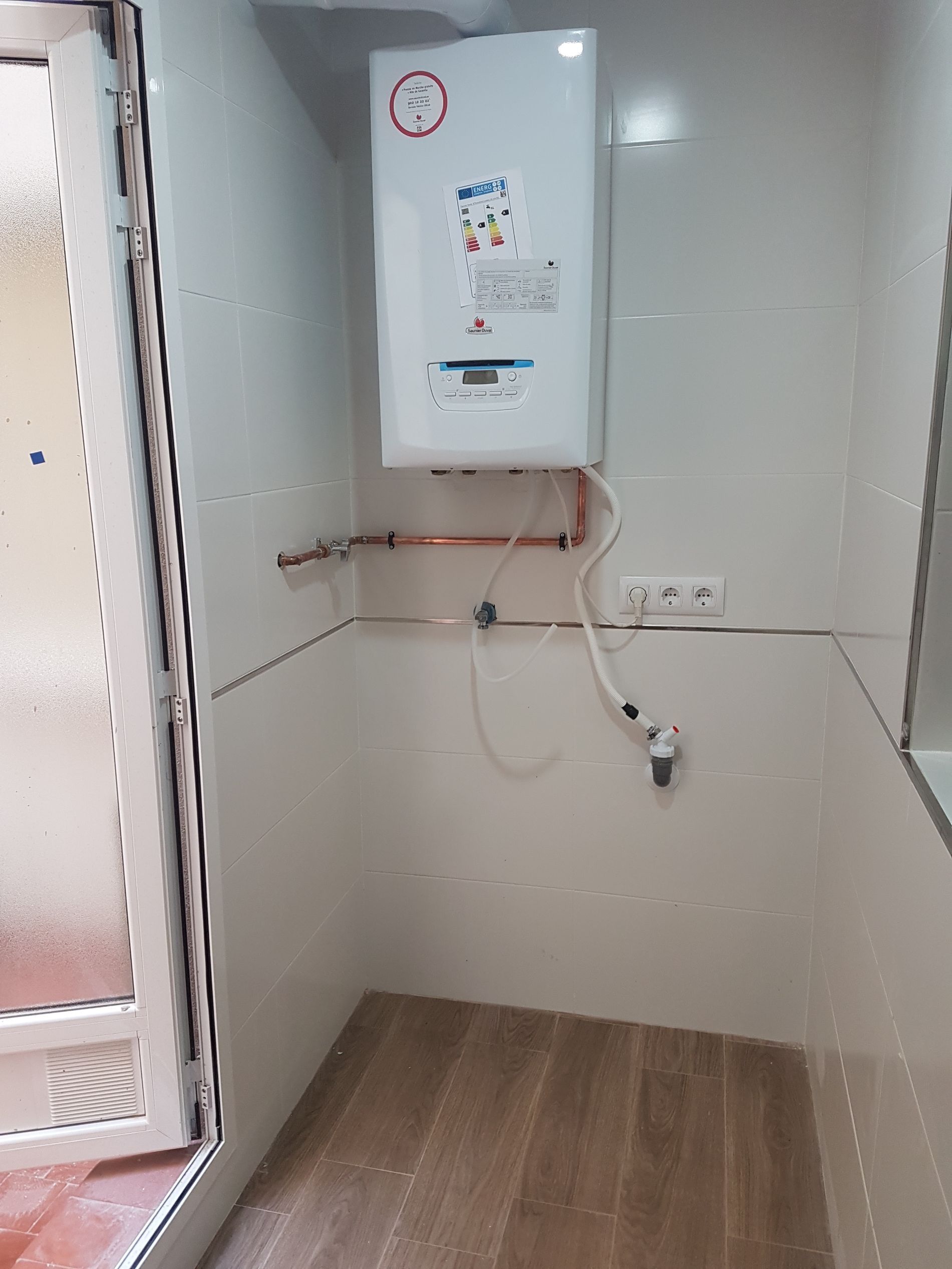 Baño pequeño con calentador de agua en la pared. Paredes de azulejos, suelo de madera y una puerta a la izquierda.