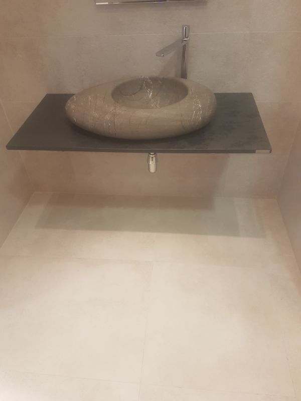 Baño con lavabo tipo piedra sobre repisa flotante gris, grifería cromada, paredes y suelo de color beige.