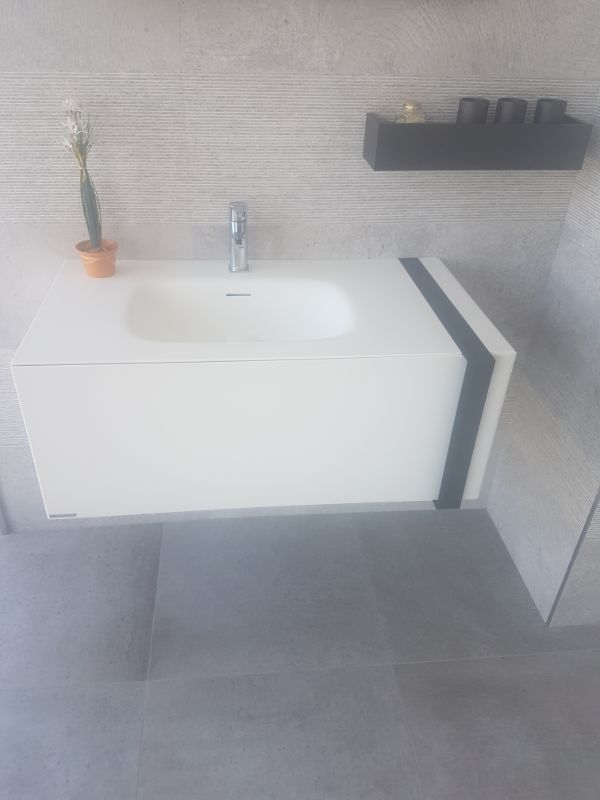 Lavabo de baño flotante de color blanco con raya negra, un pequeño cactus y un estante negro con tazas contra una pared de azulejos de color gris claro.