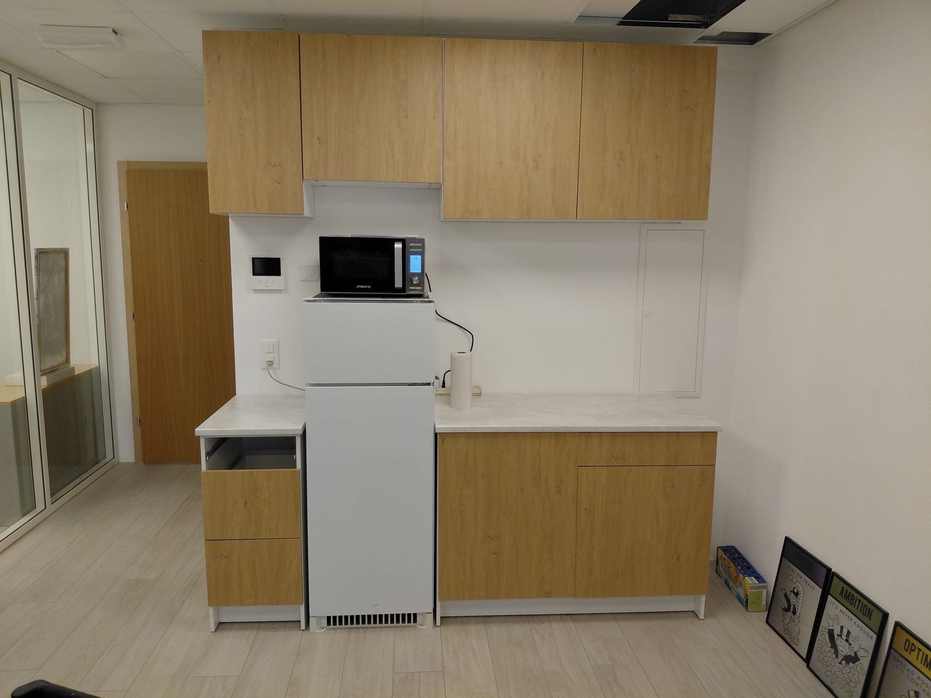 Une kitchenette dans un espace de bureau, comprenant des placards en bois clair, un réfrigérateur blanc et un four à micro-ondes.