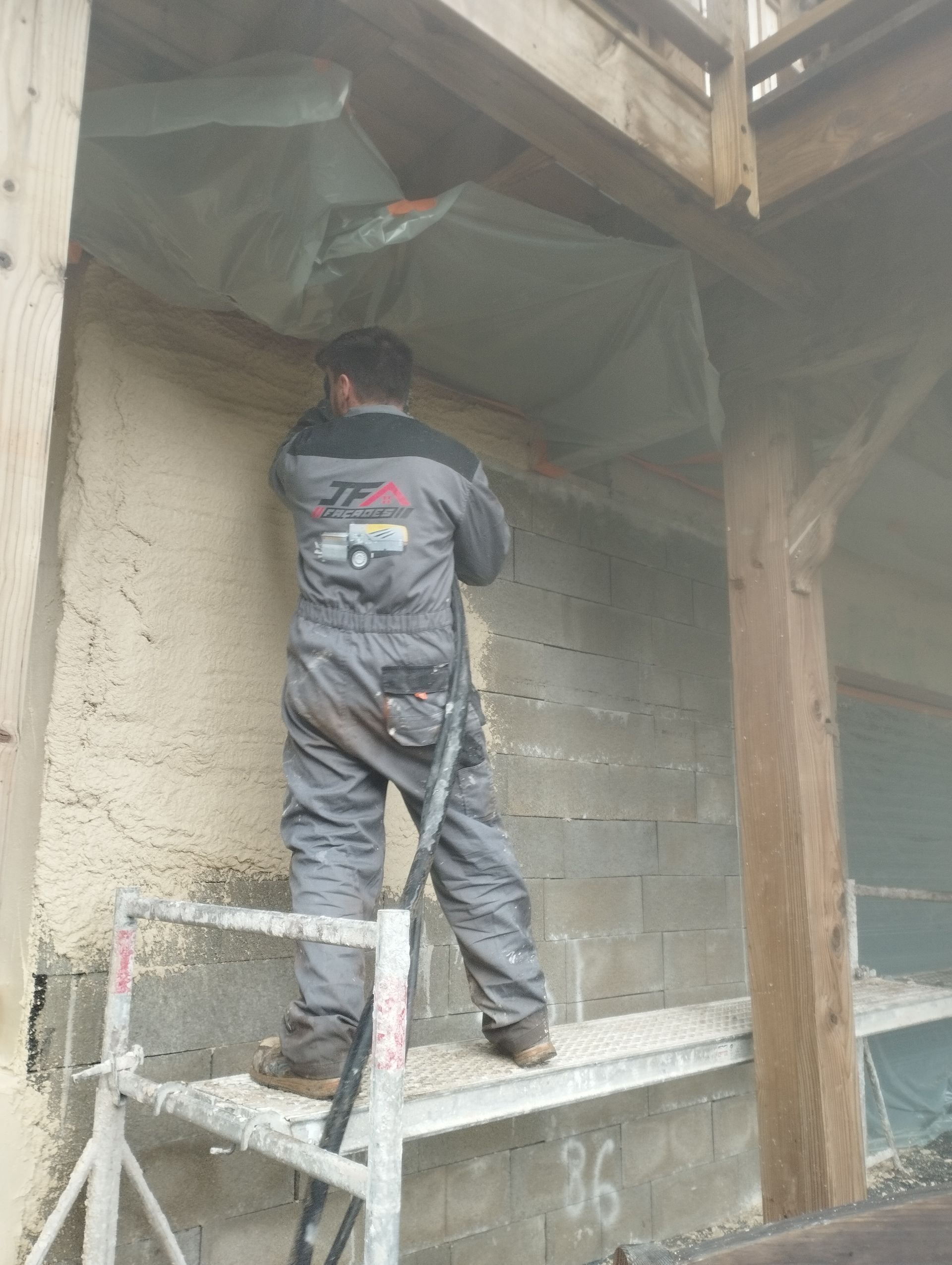 Un ouvrier du bâtiment en uniforme gris se tient sur un échafaudage, appliquant de l'enduit