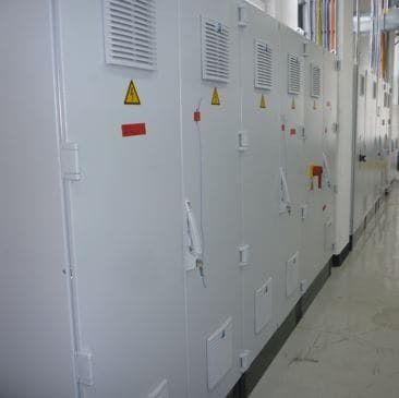 Eine Reihe weißer elektrischer Schaltschränke in einem Industriekorridor, mit Warnschildern und Lüftungsöffnungen.