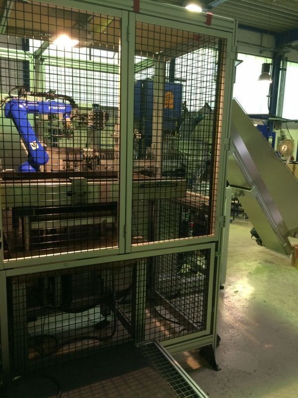 Ein blauer Industrieroboterarm in einem metallenen Sicherheitskäfig in einer Fabrik, daneben ein Förderband.