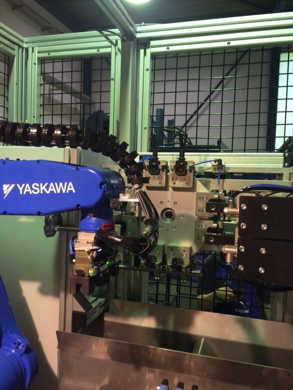 Ein Yaskawa-Industrieroboterarm arbeitet in einer umzäunten Fabrikzelle mit Metallmaschinen und Gitterfenstern.