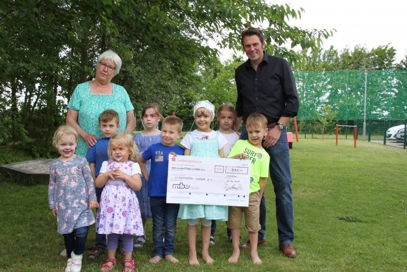 Eine Gruppe Kinder und zwei Erwachsene halten im Freien auf einem grasbewachsenen Feld einen großen Scheck.