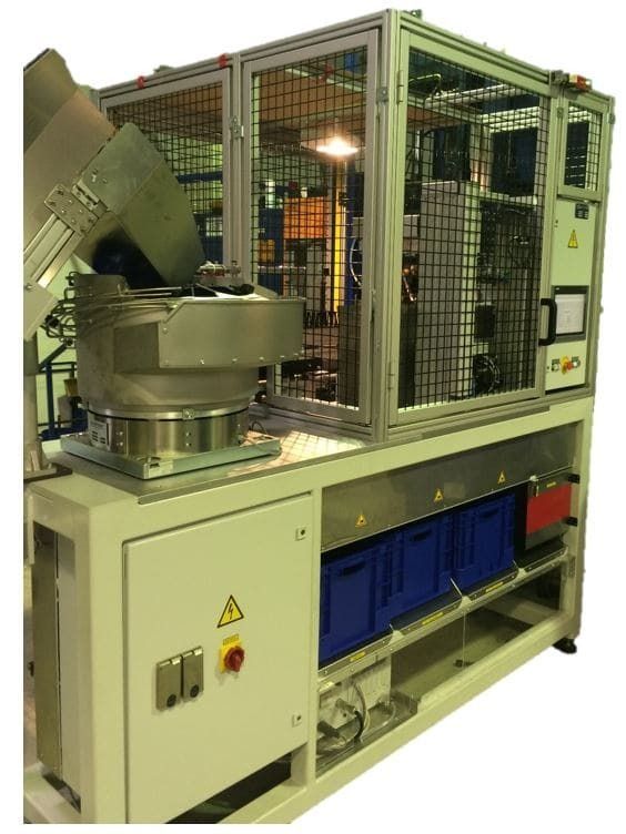 Industriemaschine mit Sicherheitskäfig, Schaltschrank und blauen Lagerbehältern in einer Fabrikumgebung