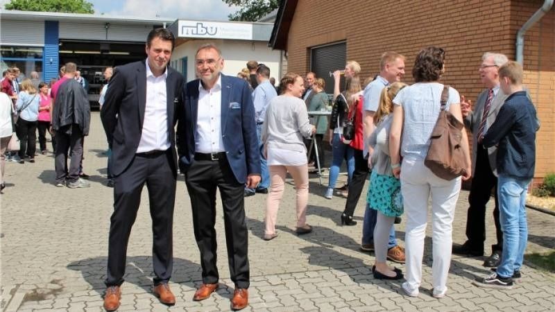 Zwei Männer in Anzügen stehen lächelnd bei einer Versammlung im Freien vor einem Backsteingebäude.