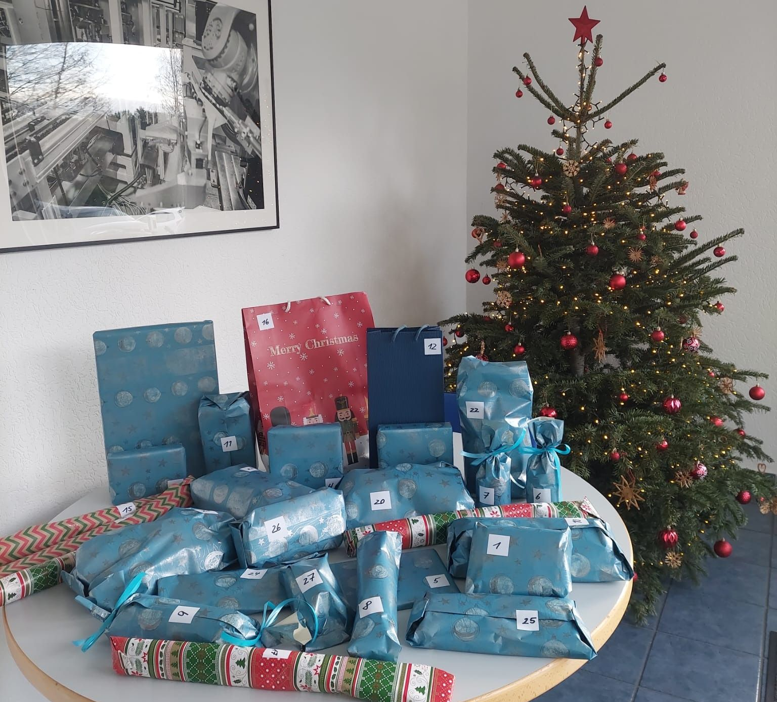 Ein Weihnachtsbaum neben einem Tisch voller verpackter Geschenke und Geschenktüten in einem hellen Raum