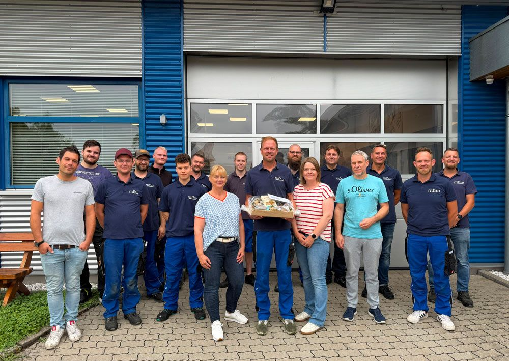 Eine Gruppe von Menschen posiert vor einem Gebäude, einer von ihnen hält ein Tablett mit Kuchen, vor einem Garagentor.