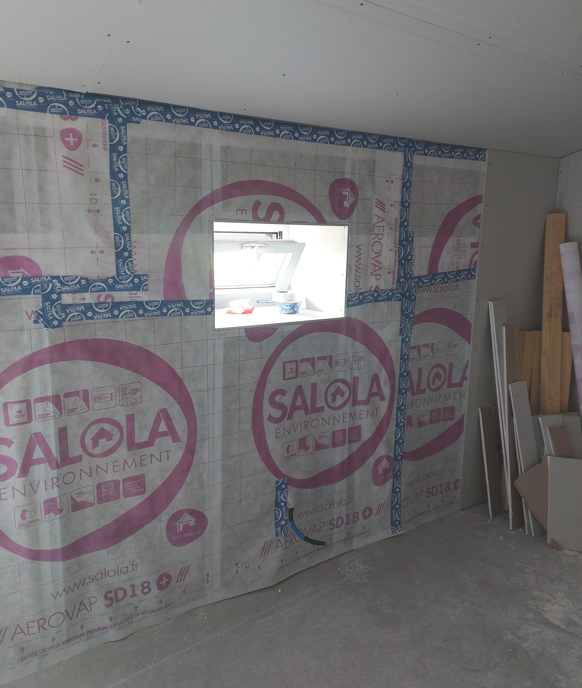 Mur recouvert d'isolant étiqueté rose, recouvert de ruban adhésif bleu. Une ouverture carrée, laisse passer la lumière du jour.