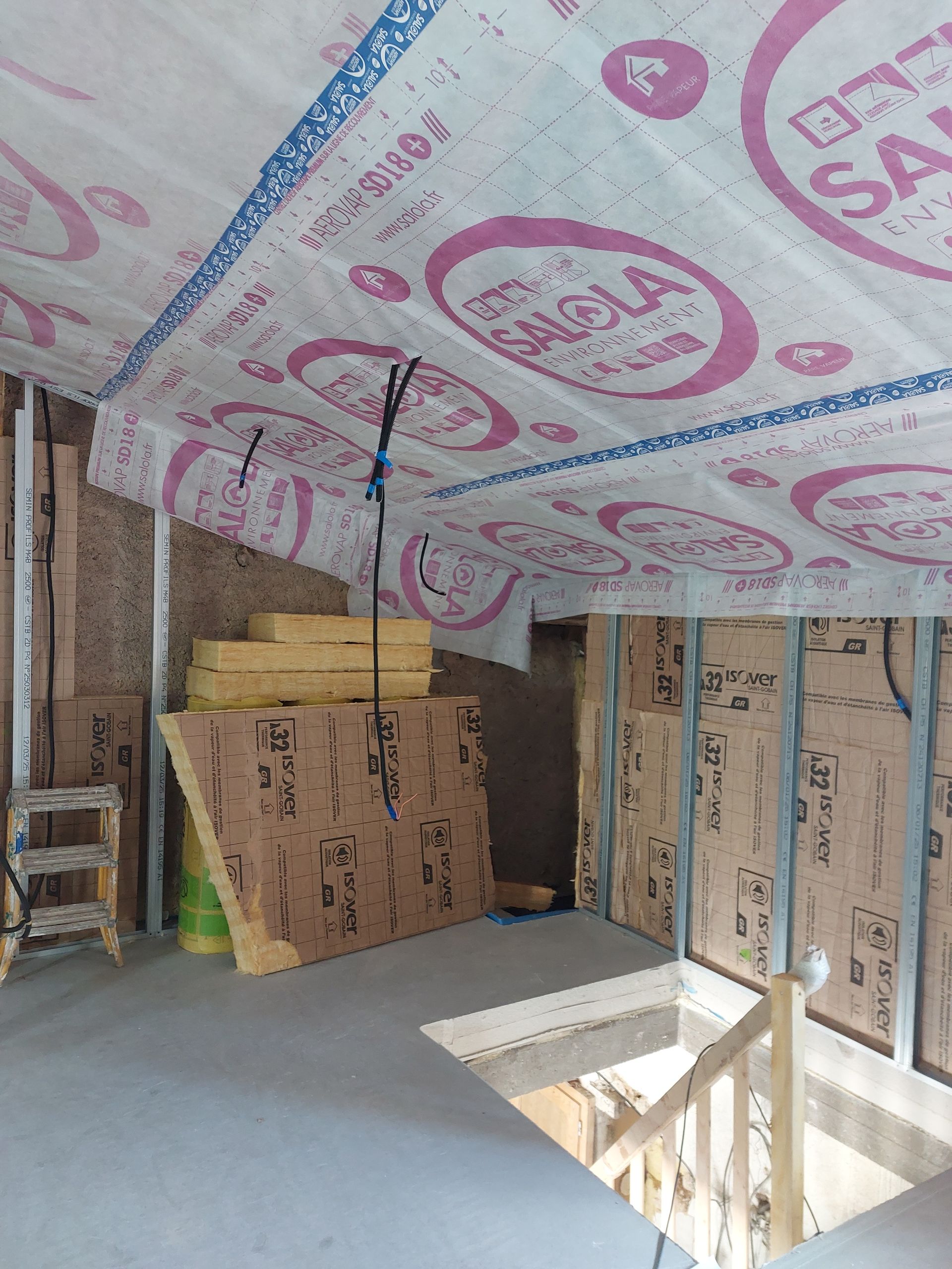 Intérieur d'une pièce en construction avec isolation apparente au plafond et aux murs ; escalier visible.