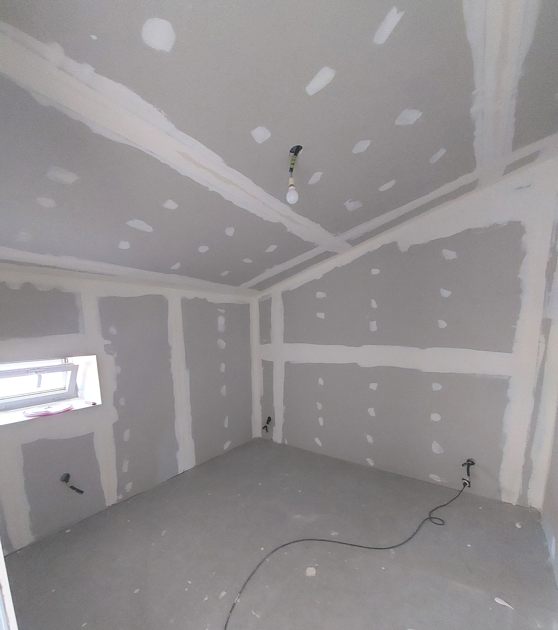 Intérieur d'une pièce en construction, murs et plafond en plaques de plâtre grises non peintes. Fils électriques apparents.