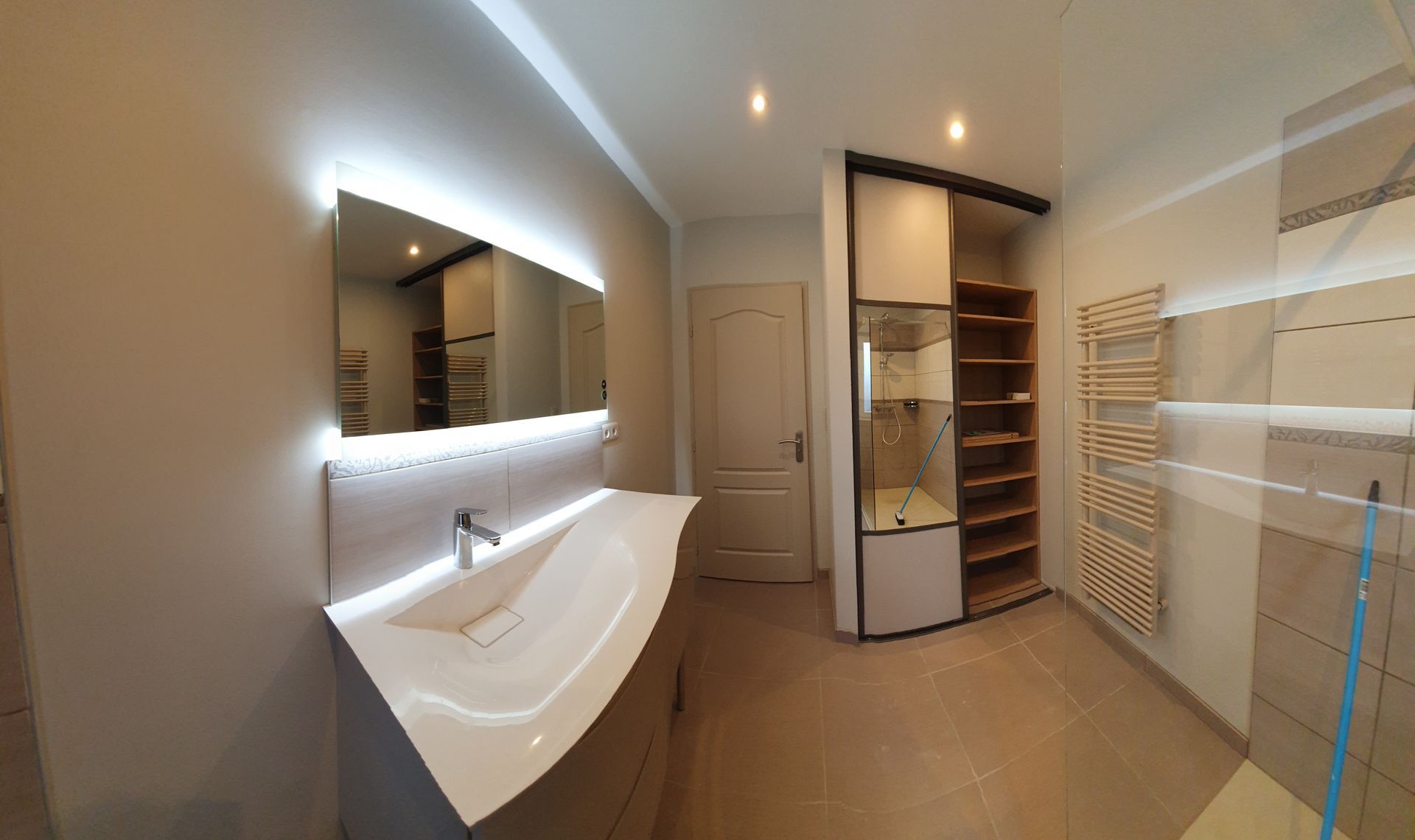 Salle de bains moderne avec lavabo blanc, miroir éclairé, rangements en bois et douche.