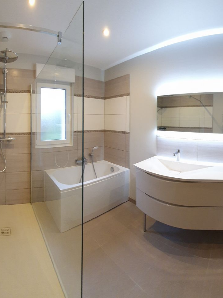 Salle de bains moderne avec baignoire, douche et meuble pour lavabo. Murs carrelés beiges et blancs et sol clair.